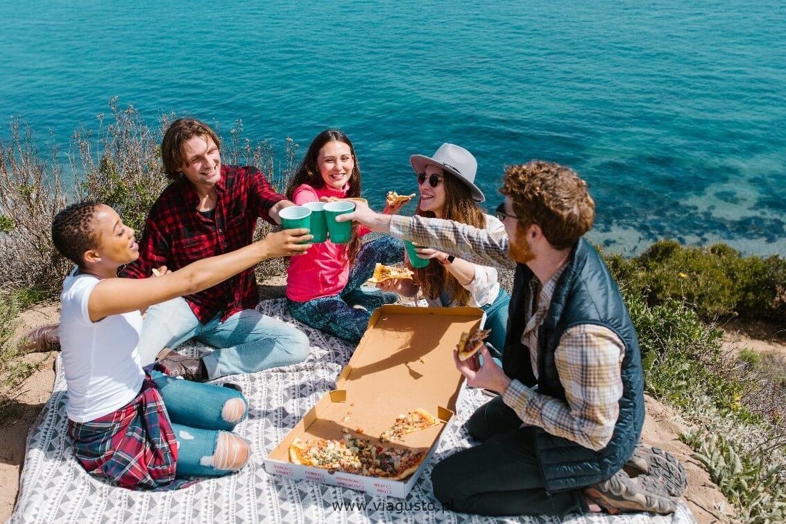 mężczyźni i kobiety wznoszą toast na biało niebieskim kocu, na którym leży pudełko pizzy